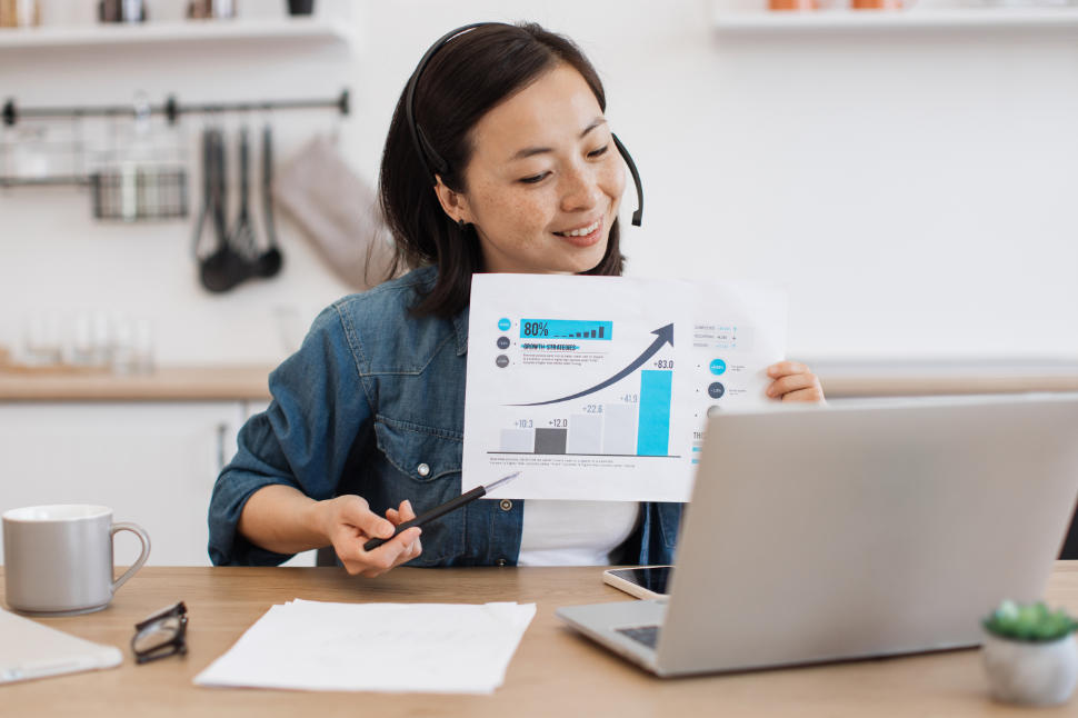 Cheerful young lady in headset pointing at graphs on paper report while holding online briefing via laptop at home. Efficient female expert interpreting current data for making business predictions.