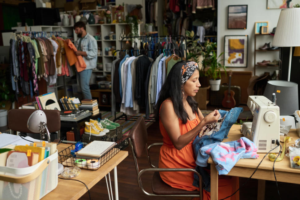 Young woman in casualwear sitting by table with electric sewing machine and decorating denim jacket with beads against customer
