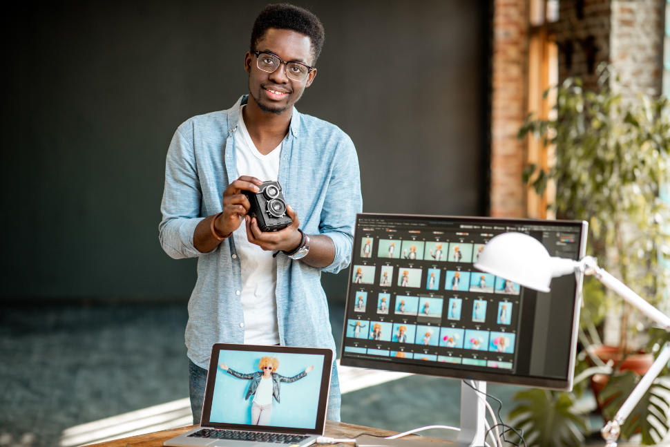 Portrait of a young african ethnicity photographer standing with retro camera near the working place with computers in the studio
