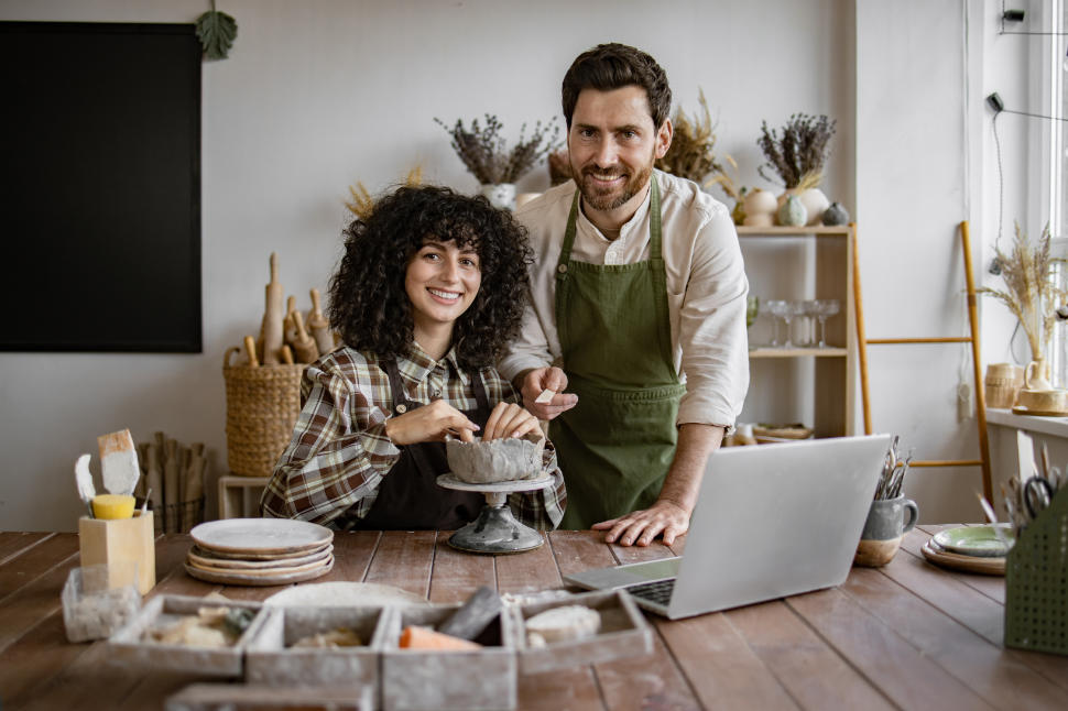 Smiling couple working together on pottery project in art studio. Creative process with laptop and ceramic tools on wooden table.