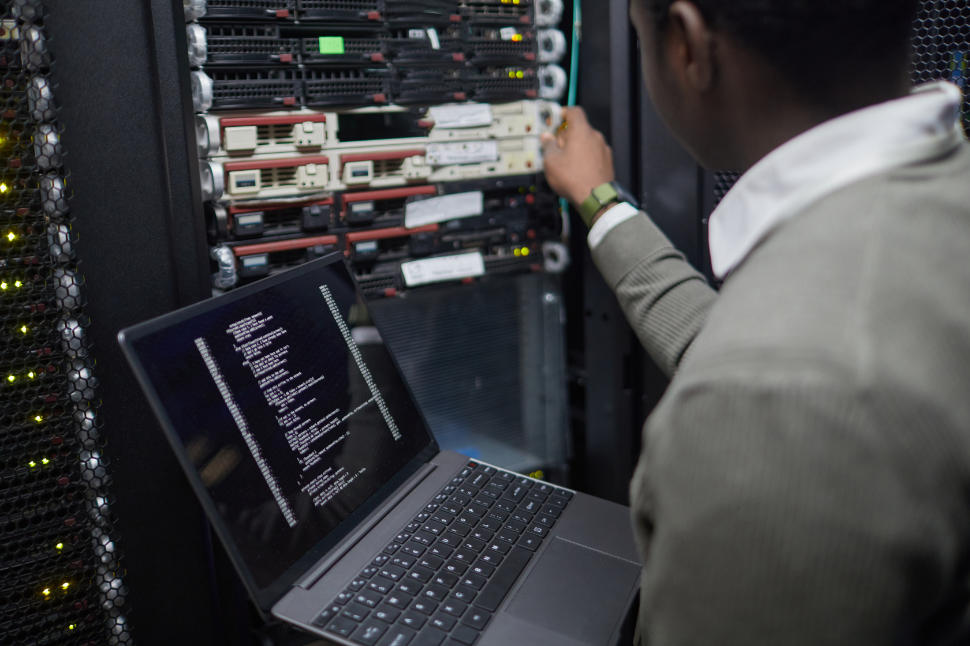 Person managing server equipment in a technology environment, checking system status on a laptop. Focus on information technology and network administration