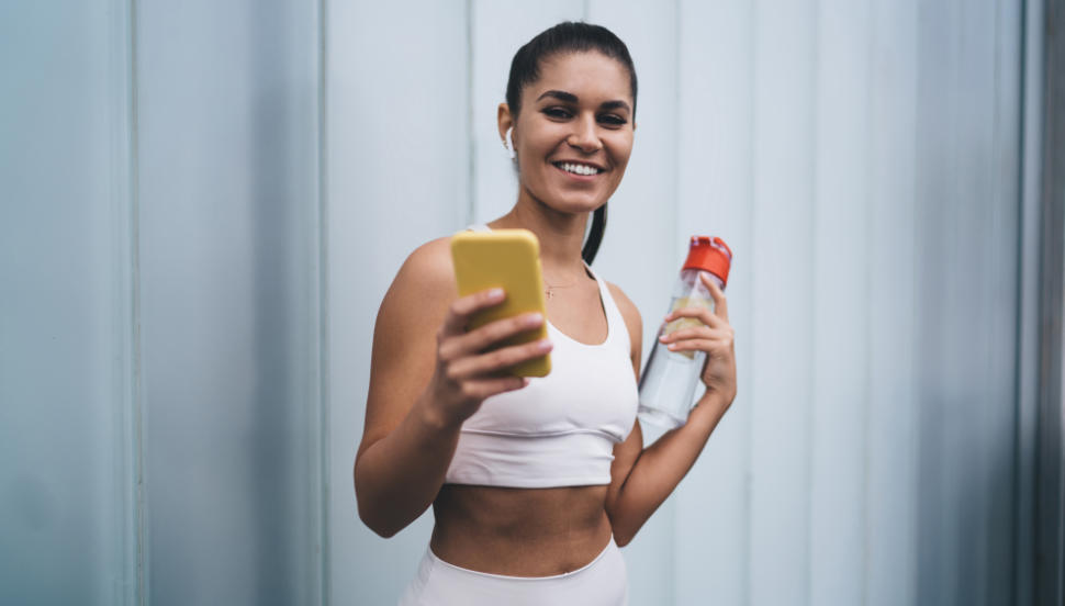 Portrait of young smiling female in wireless earphones standing with mobile phone device on copy space background. Attractive brunette woman with fit figure controls physical activity using mobile app