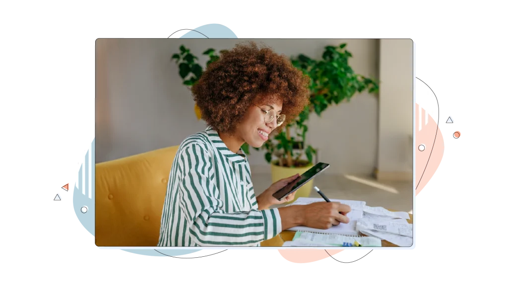 Woman smiling and writing notes on her notebook while using her smartphone in her office.