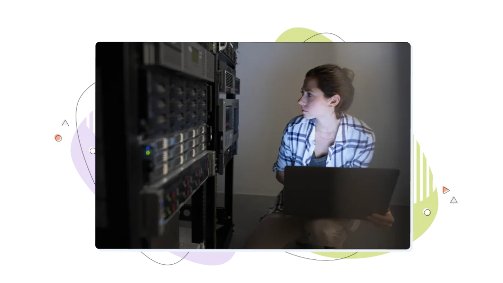 Woman typing on her laptop in a server room, while looking at the server.