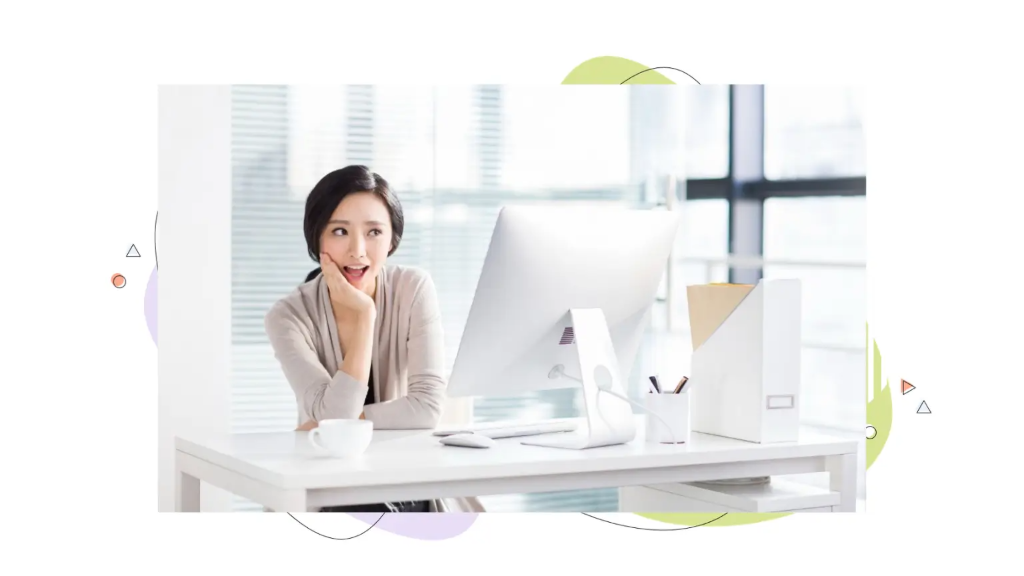 Woman sitting at a bright office desk looking surprised and delighted at her desktop monitor while discovering the benefits of VPS hosting.